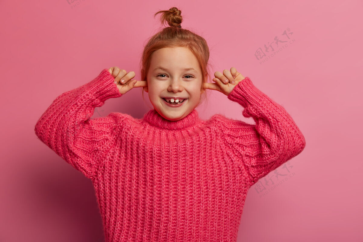 搞笑可爱快乐的小女孩用食指塞住耳朵 开心地笑出来 不理会响亮的声音 留着发髻 穿着针织毛衣 在玫瑰色的墙上摆姿势我听不见你声音室内积极
