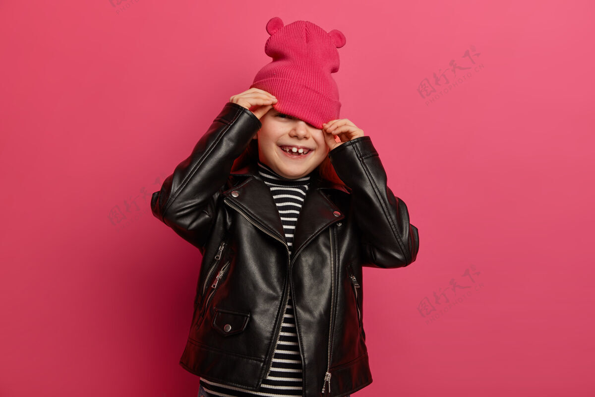 欢乐快乐的欧洲女孩戴着带耳朵的粉色帽子 黑色皮夹克 在妈妈给她拍照时躲起来 表情欢快 在粉色充满活力的墙上摆姿势风格和童年欢呼积极寻求