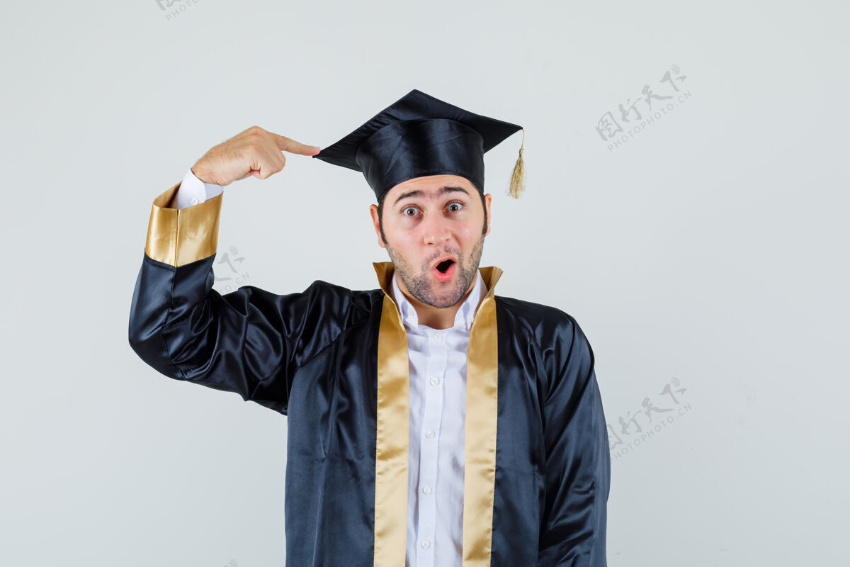 休闲年轻人指着他那顶穿着毕业制服的黑帽子 惊讶地看着 正前方的景色男人年轻自信