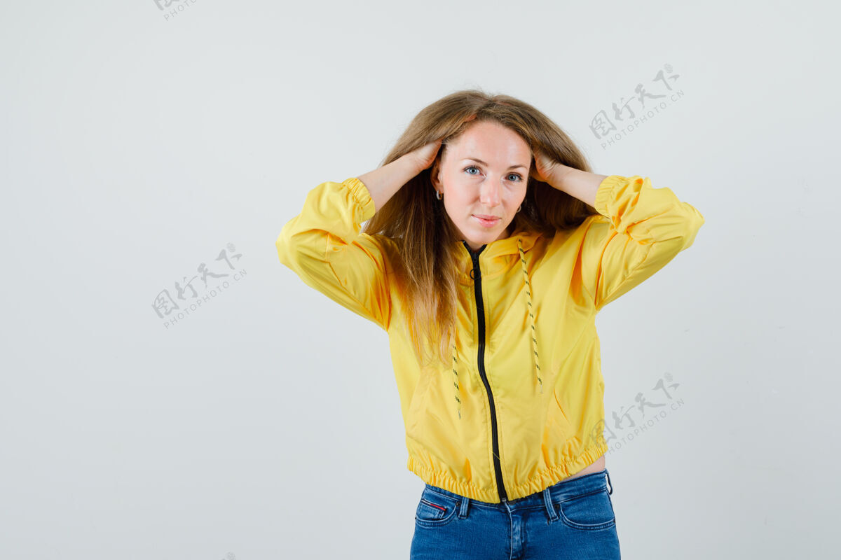 女性身穿黄色棒球服和蓝色牛仔裤的年轻女子手放在头上 看起来很有吸引力 正面视图成人年轻夹克