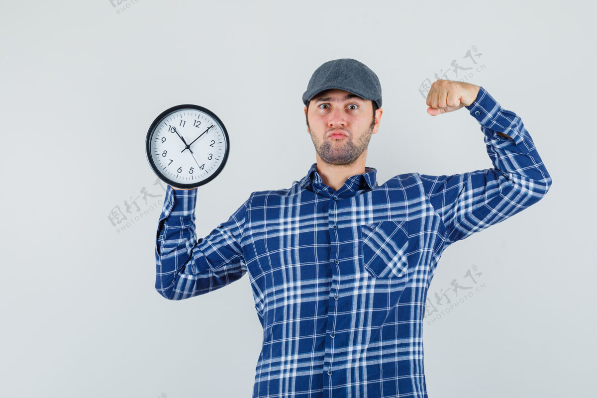 男人年轻人拿着挂钟 展示着衬衫 帽子上的肌肉 看上去很自信正视图时钟聪明男性