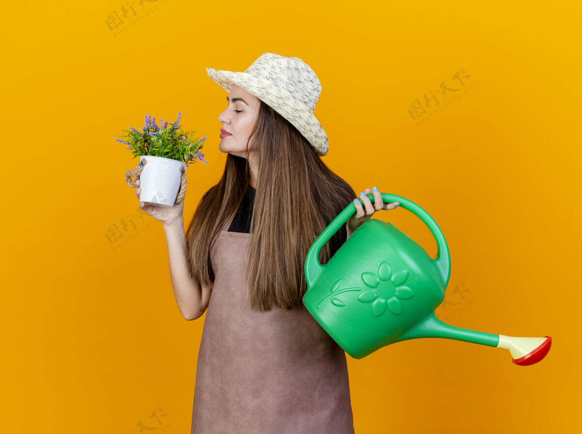 请高兴美丽的园丁女孩穿着制服 戴着园艺帽 手里拿着浇水罐 嗅着花盆里的花 孤立在橙色的背景上浇水手帽子