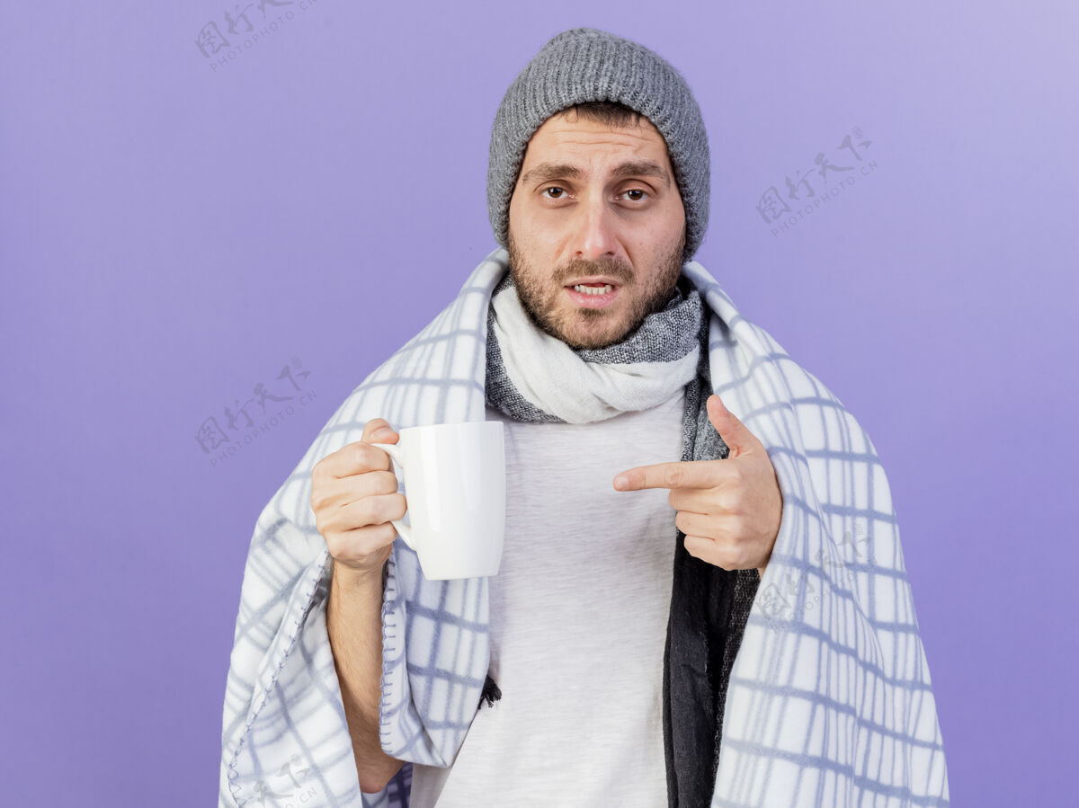 年轻一个戴着冬天帽子 戴着围巾 指着紫色背景上孤立的一杯茶的年轻病患围巾举行茶