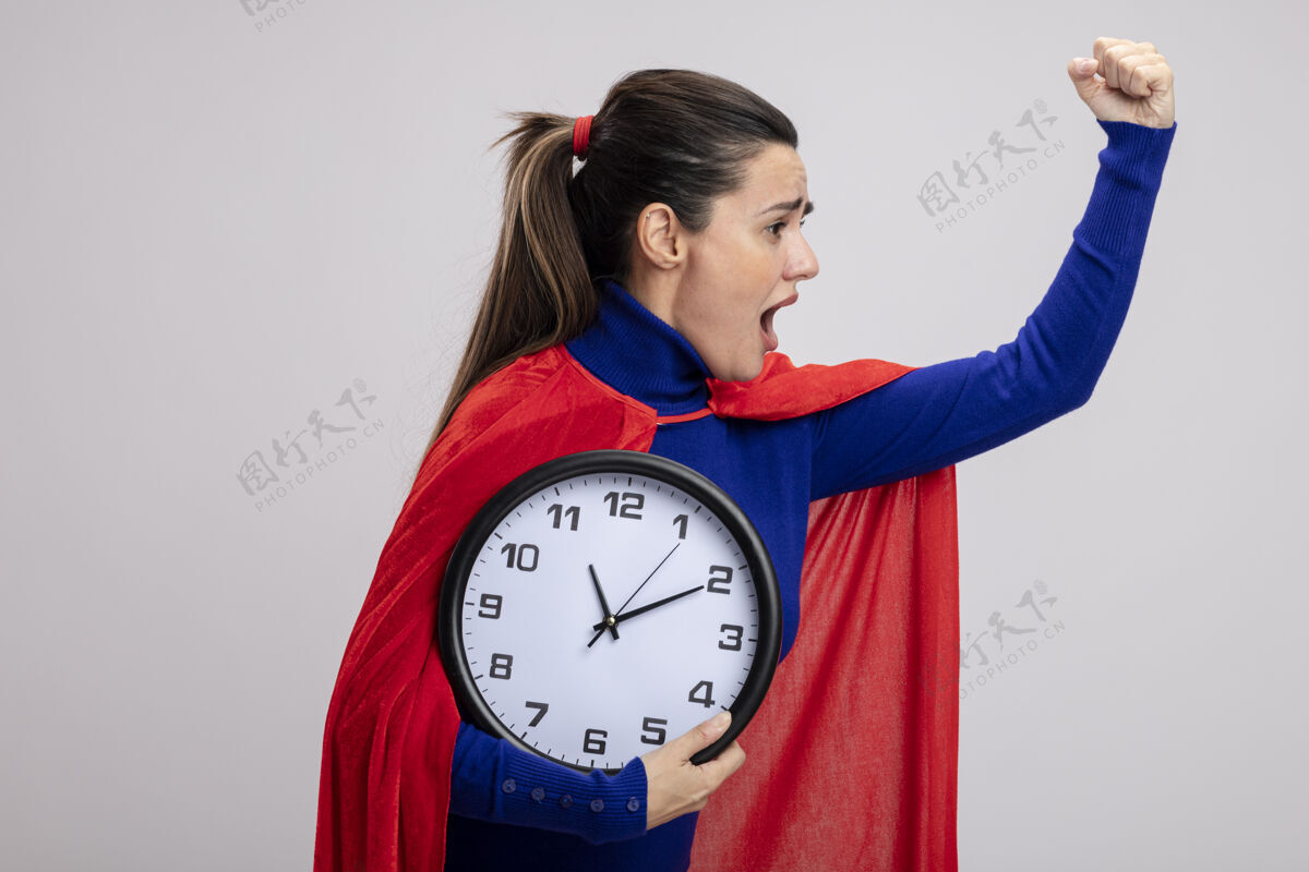 举起愤怒的年轻超级英雄女孩站在侧视图中举着挂钟 举起手孤立在白色背景上站着抱着手