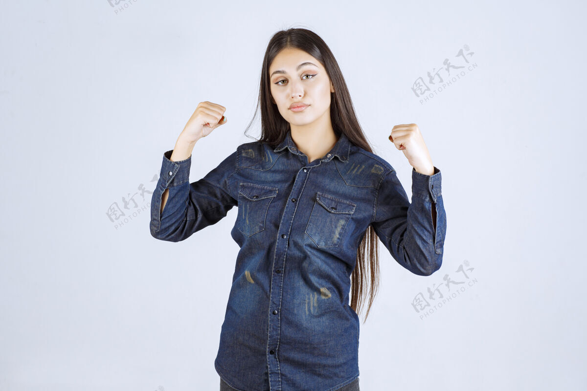 运动穿着牛仔衬衫的年轻女子展示了她的手臂肌肉人工人拳头