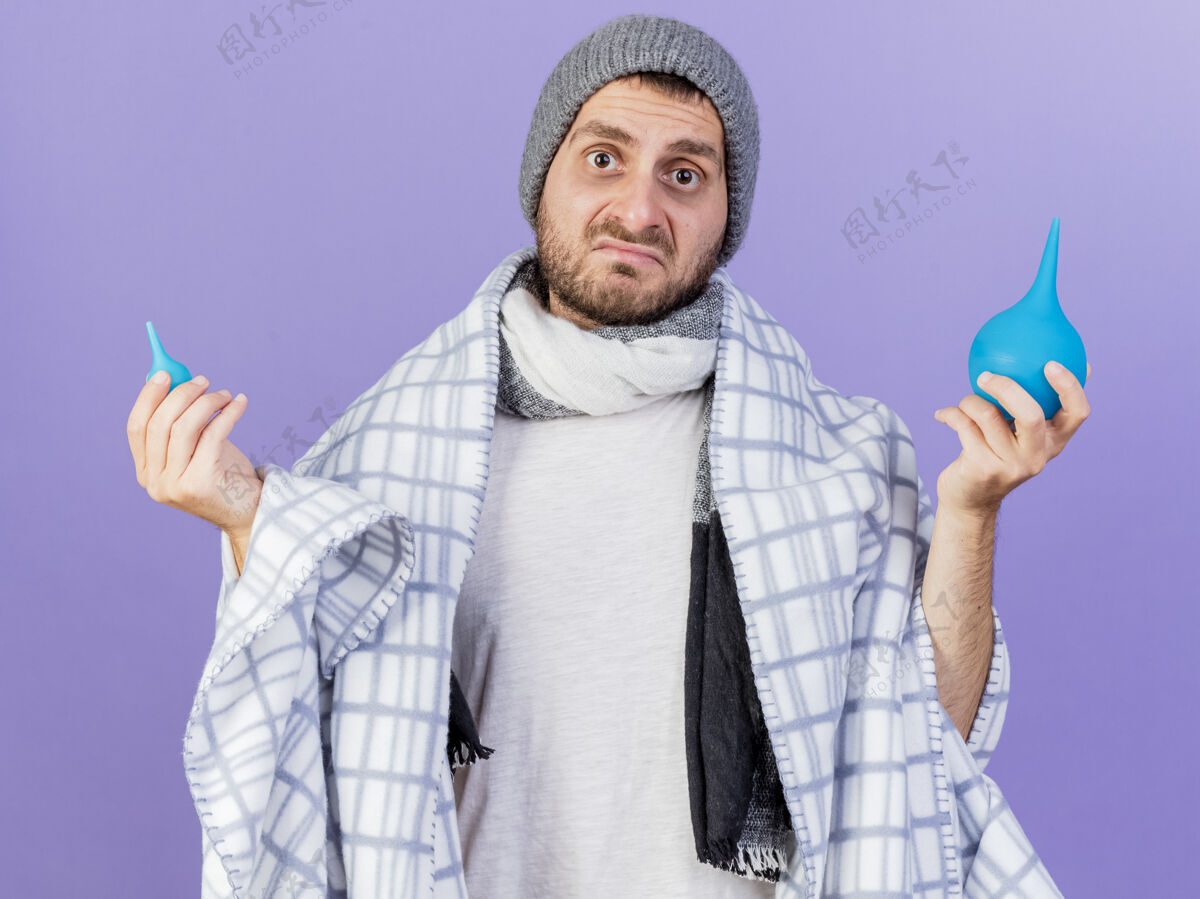 裹着困惑的年轻病人戴着冬天的帽子 围巾裹着格子布 拿着灌肠剂 摊开双手孤立在紫色背景上摊着迷糊年轻