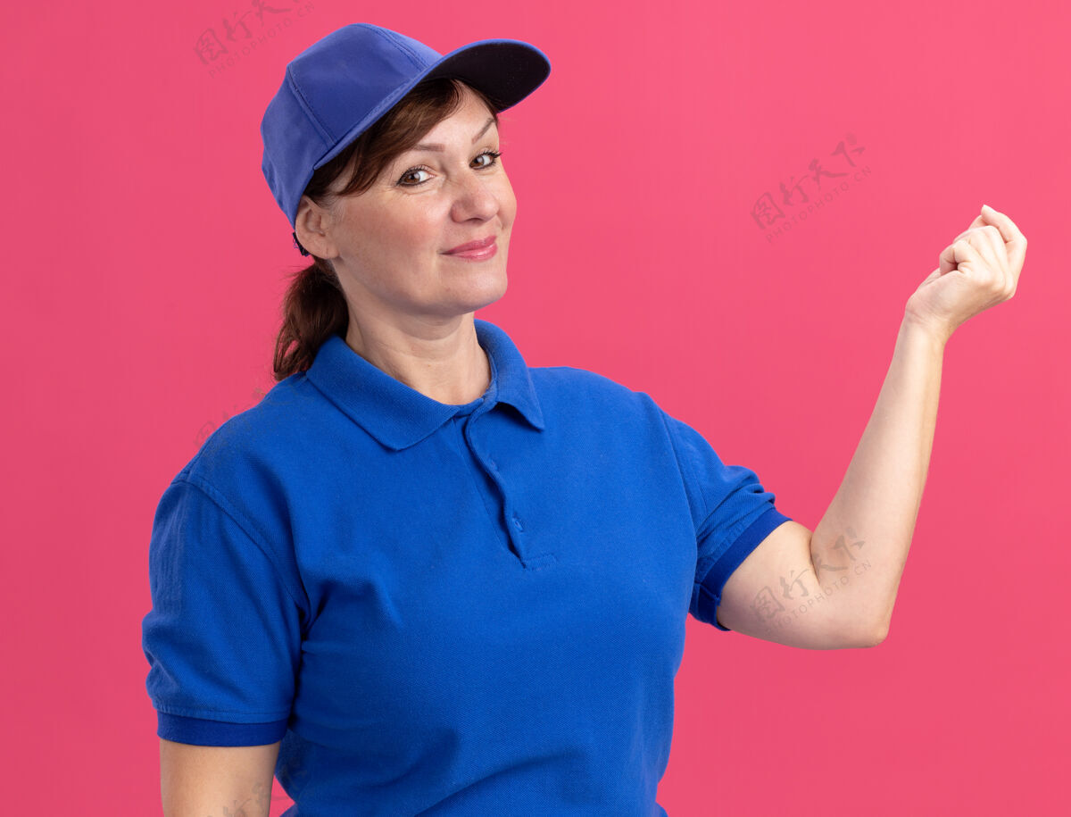 钱身穿蓝色制服 头戴帽子的中年送货妇女站在粉红色的墙上 微笑着看前面赚钱 手势是搓手指等待付款微笑付款送货