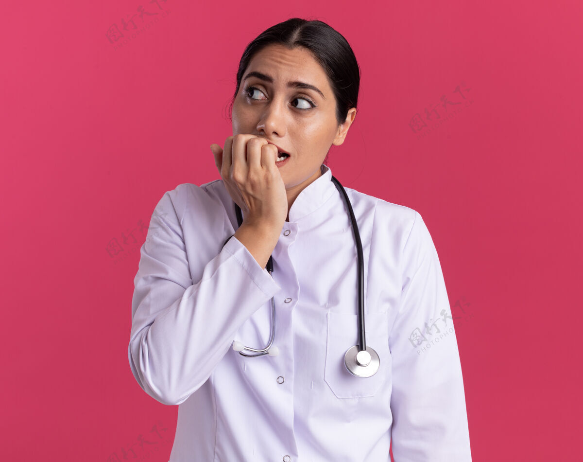 医生年轻的女医生穿着医用外套 手持听诊器 站在粉红色的墙上 紧张地看着旁边咬着的指甲年轻人女人靠边站