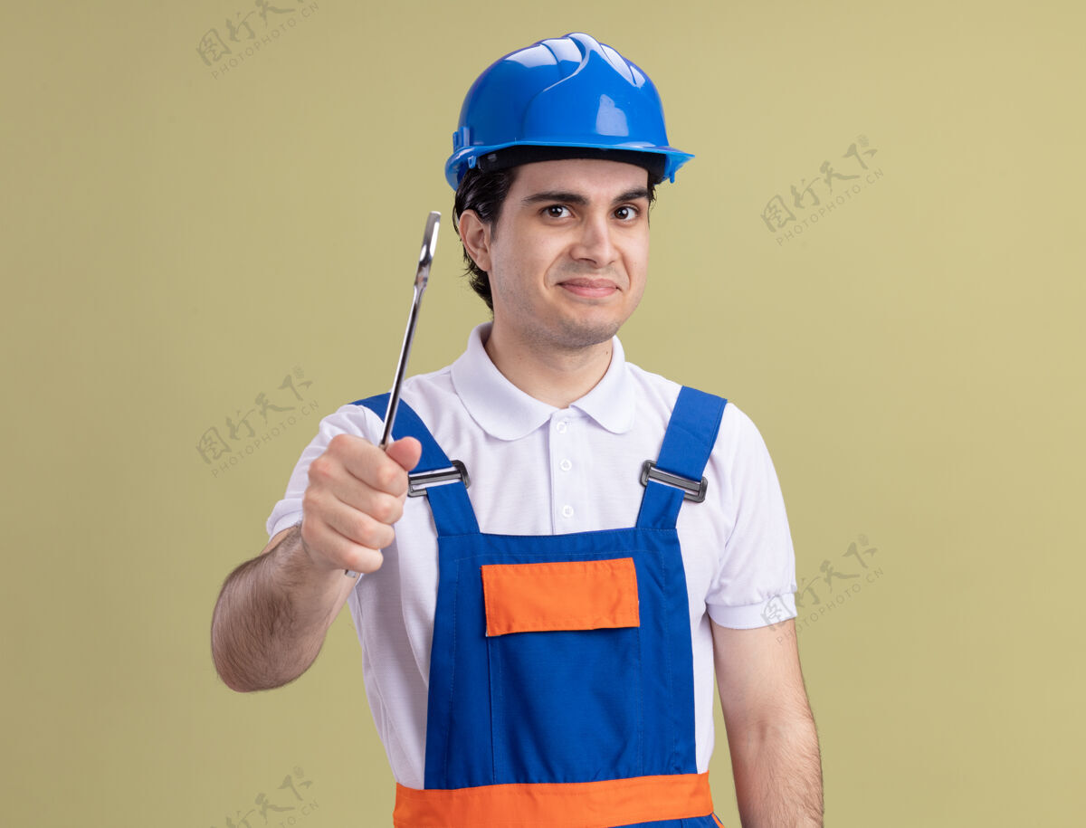 建筑年轻的建筑工人穿着建筑制服 戴着安全帽 拿着扳手 站在绿色的墙上微笑着看着前面站着头盔建筑工人