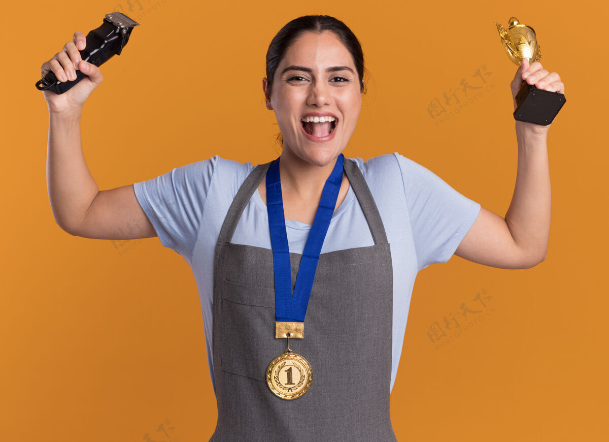举起年轻漂亮的女理发师围着围裙 脖子上挂着金牌 手里拿着修剪器和金杯 高兴而兴奋地站在橙色的墙上女人金牌奖牌