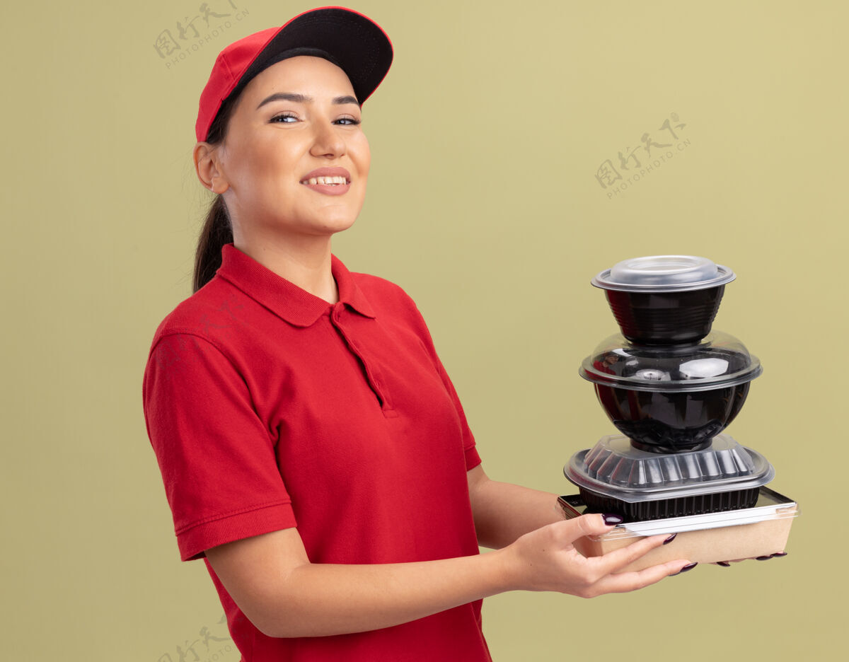 快乐身着红色制服 戴着帽子的快乐的年轻女送货员 手里拿着一叠食品包 面带微笑地站在绿色的墙边看着前方站着年轻制服