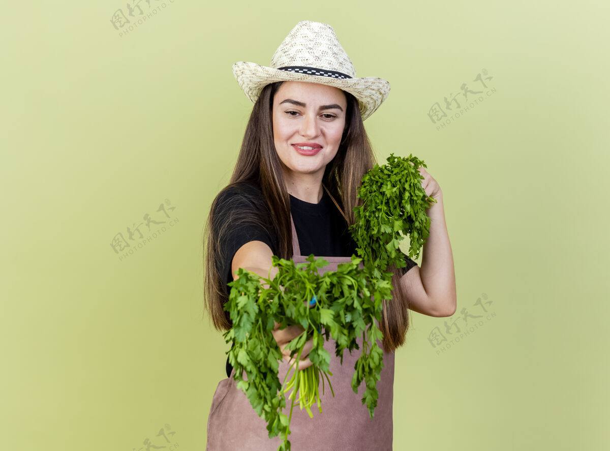 花园身着制服 面带微笑的美丽园丁女孩戴着园艺帽 手持芫荽叶 对着橄榄绿背景上孤立的镜头微笑香菜相机