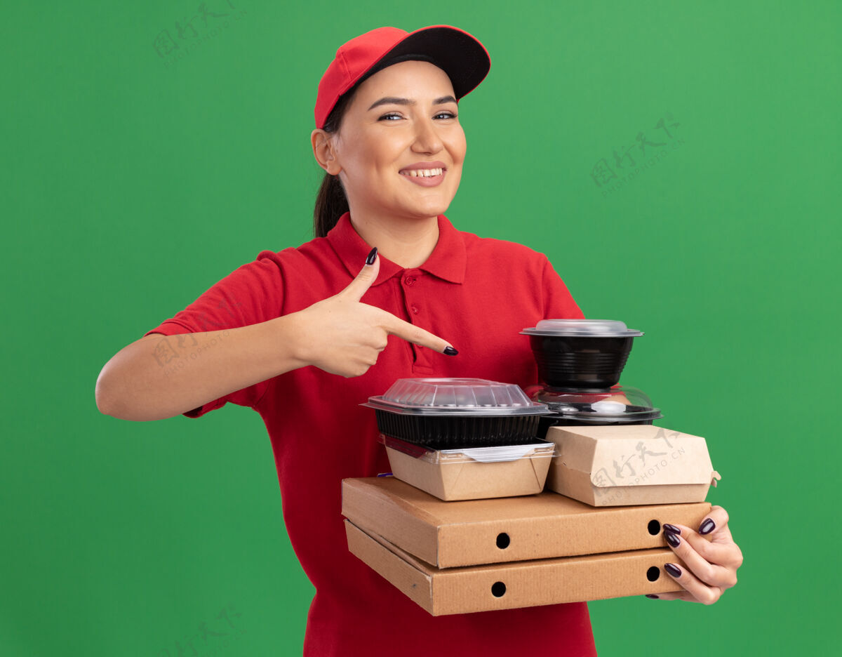 帽子身穿红色制服 头戴鸭舌帽的年轻女送货员拿着披萨盒和食品包 用食指指着他们 站在绿色的墙边开心地微笑着站着指着目录