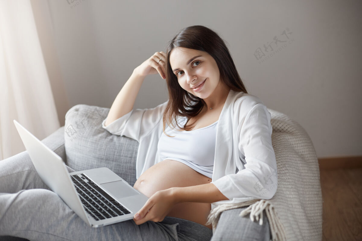 女孩孕妇用笔记本电脑坐在家里舒适的椅子上为人父母成人新的