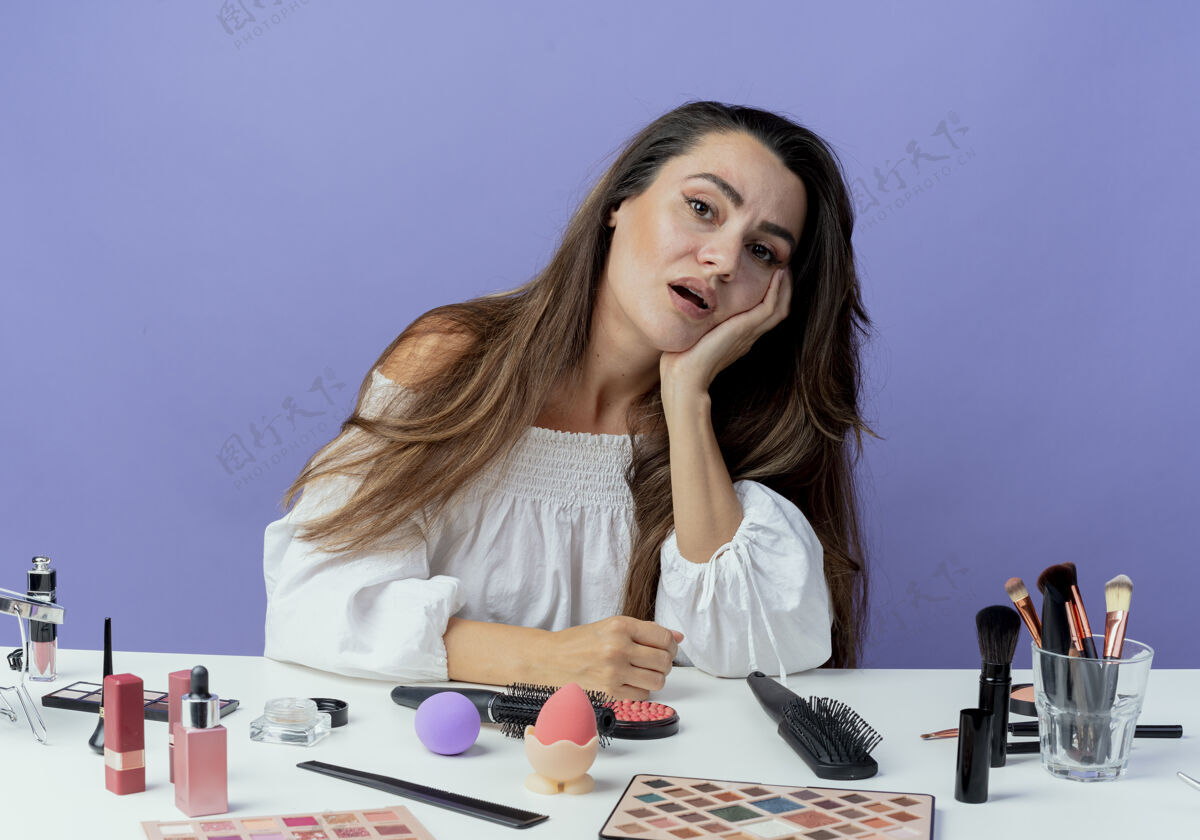 女人疲惫美丽的女孩坐在桌子旁 用化妆工具把手放在下巴上 看起来孤立在紫色的墙上工具桌子紫色
