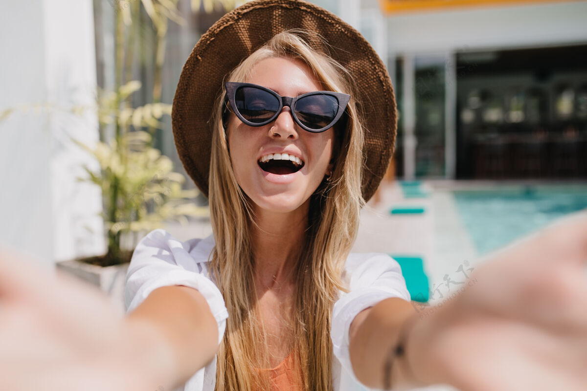 年轻戴着迷人太阳镜的金发女人表达积极的情绪兴奋的高加索女士在度假村自拍的户外镜头帽子活跃女人