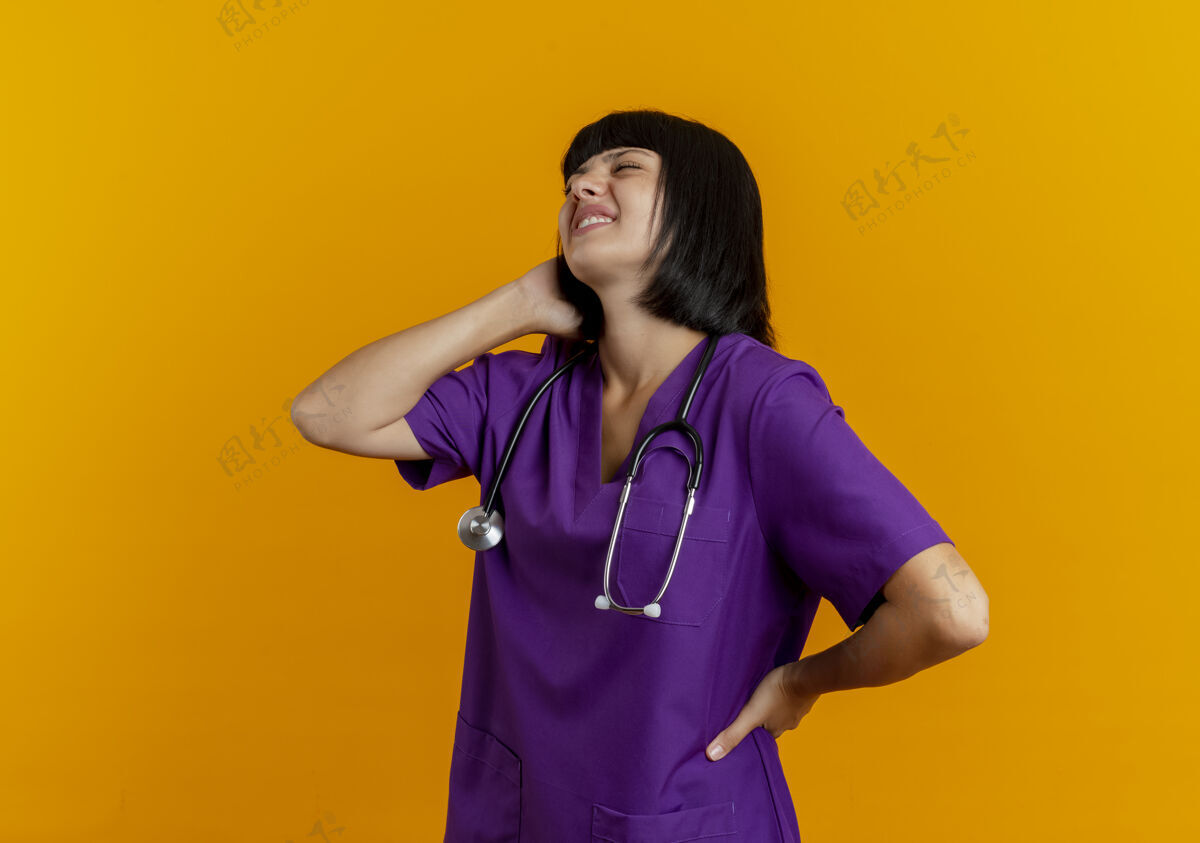 黑发疼痛的年轻黑发女医生穿着制服 手持听诊器站在一边 脖子后面拿着疼痛站着