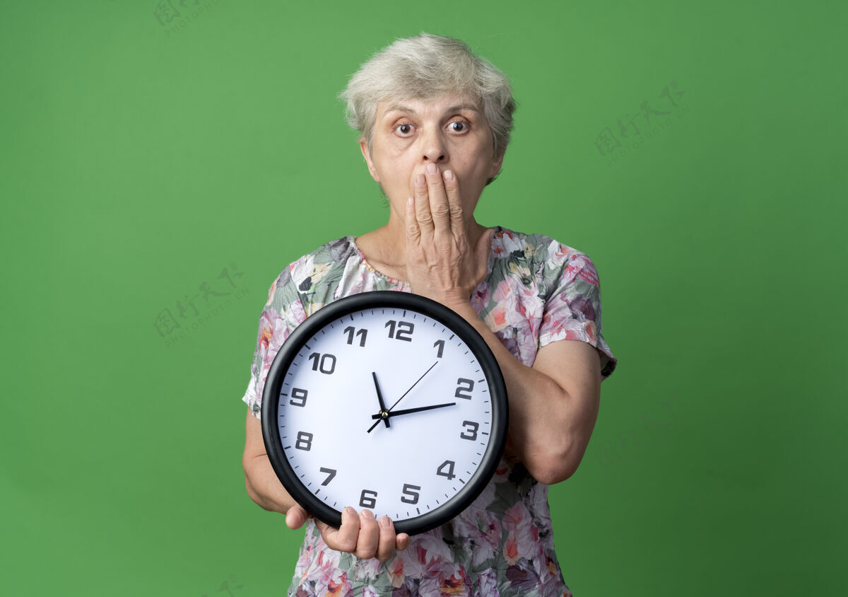 手震惊的老妇人把手放在嘴上 拿着隔离在绿墙上的钟钟嘴女人