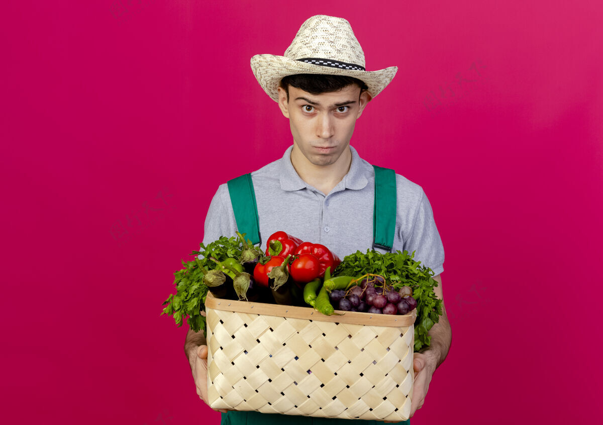 持有心烦意乱的年轻男园丁戴着园艺帽拿着菜篮子看着复制穿蔬菜