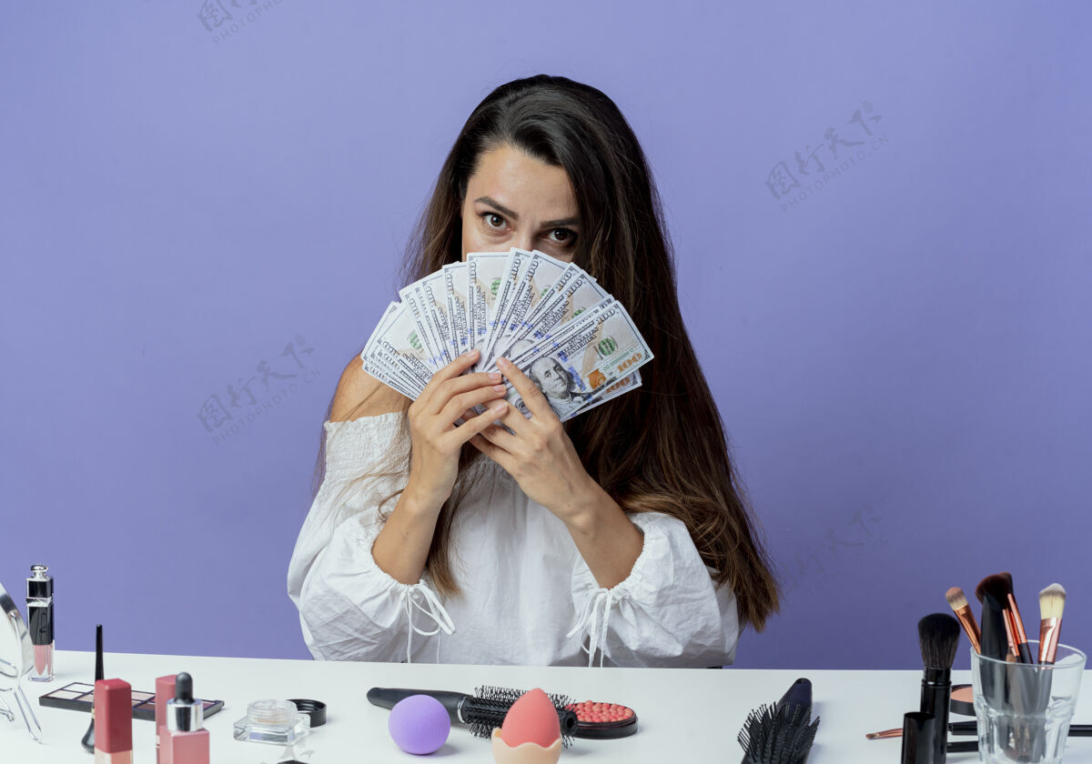 紫色惊讶的美丽女孩坐在桌子旁 拿着化妆工具 看着紫色墙上孤立的钱女人工具桌子