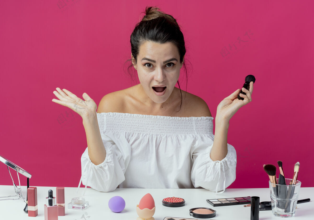 画笔惊讶美丽的女孩坐在桌旁 拿着化妆工具 举着化妆刷 双手孤立在粉红色的墙上举起女人惊喜