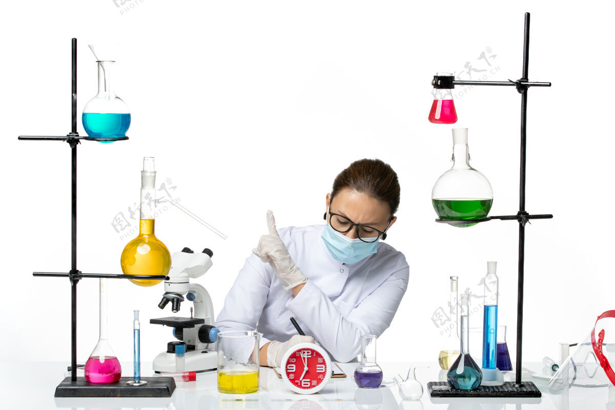 笔记前视图穿着医疗服的女化学家戴着面罩坐在那里 拿着溶液在白色背景上写笔记化学家实验室病毒covid-splash医疗容器喷溅