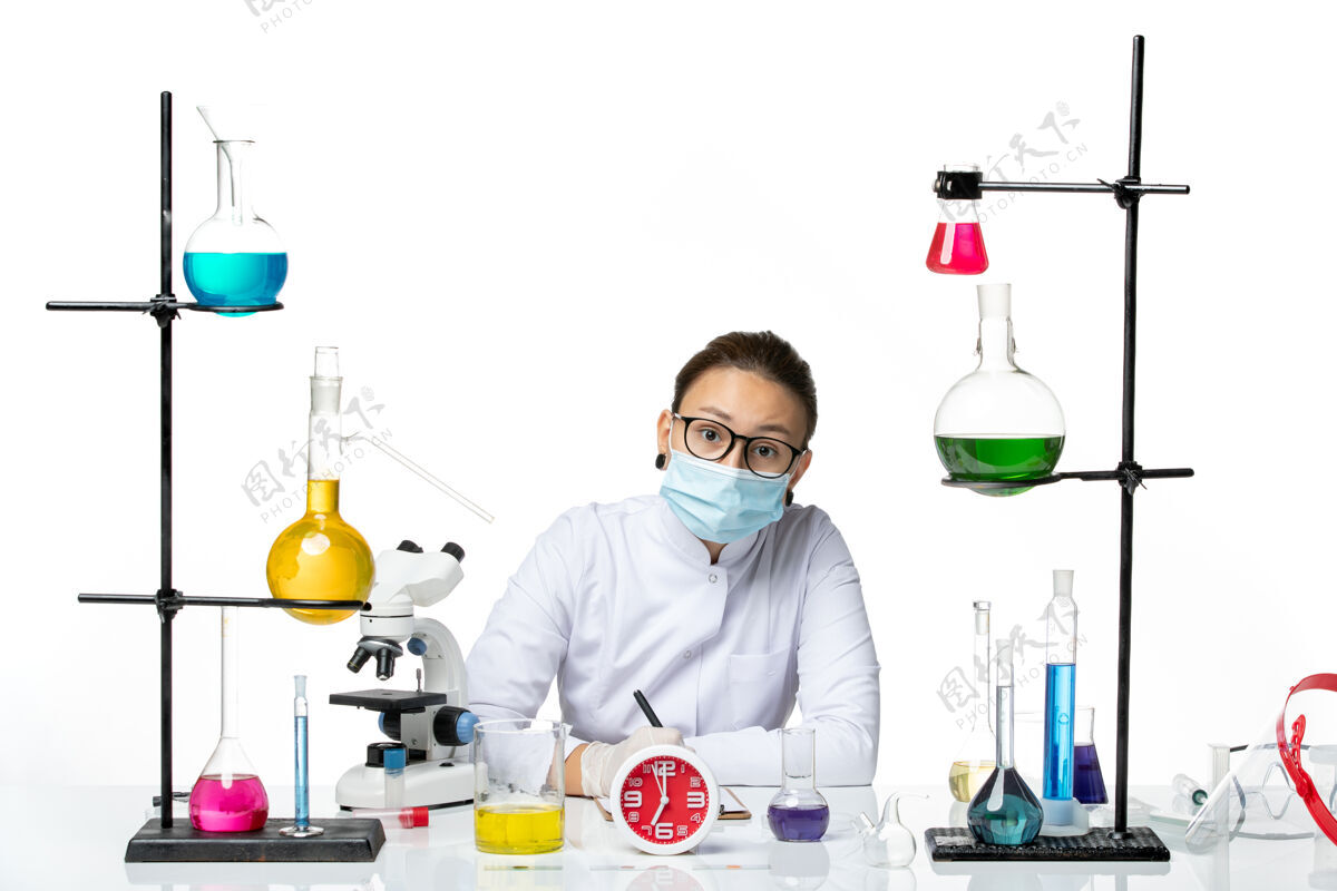 喷溅前视图穿着医疗服的女化学家戴着面罩坐在那里 拿着溶液在白色背景上写笔记化学家实验室病毒covid-飞溅医疗视图坐着