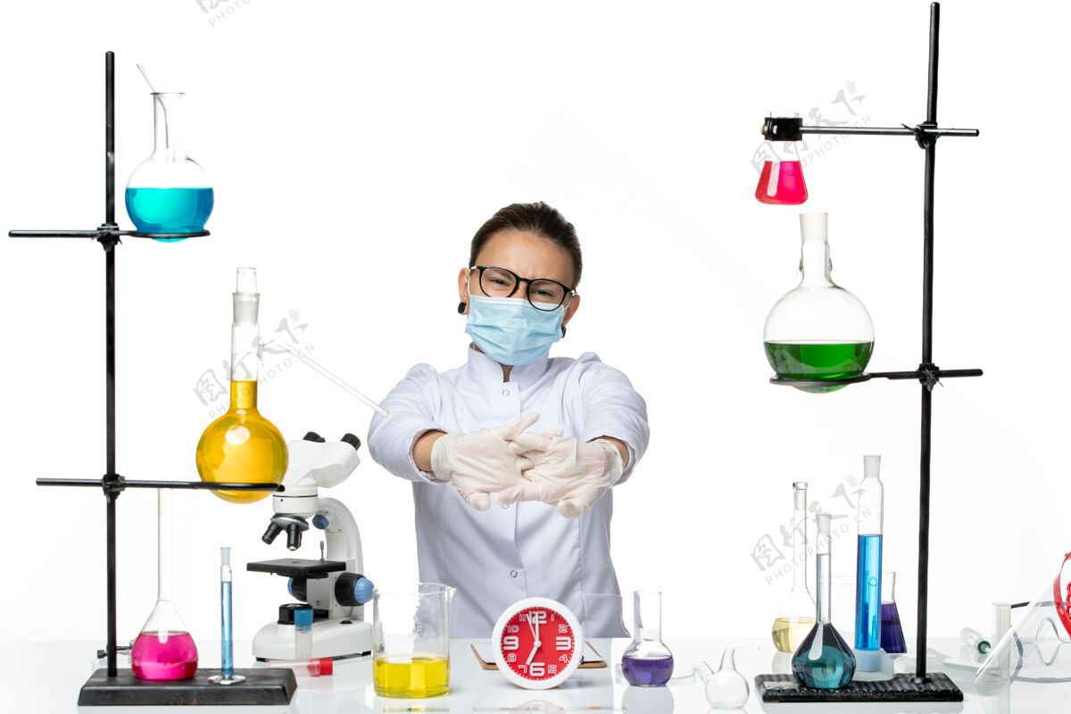 实验前视图穿着医疗服的女化学家戴着面罩坐在白色的办公桌上拿着溶液溅起病毒化学病毒-实验室套装溶液前面
