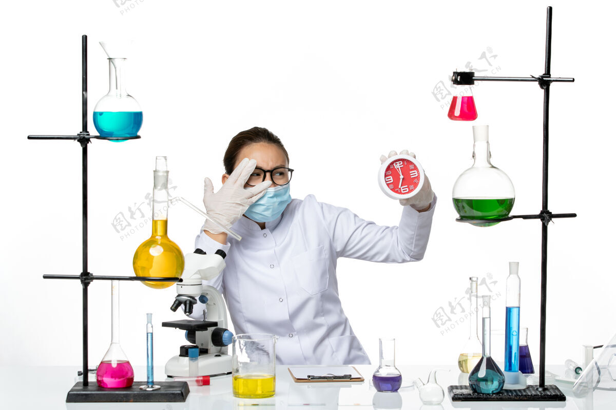医学前视图：穿着医疗服的女化学家 戴着面罩 拿着浅白色背景上的时钟病毒实验室化学冠状病毒飞溅烧杯科学时钟