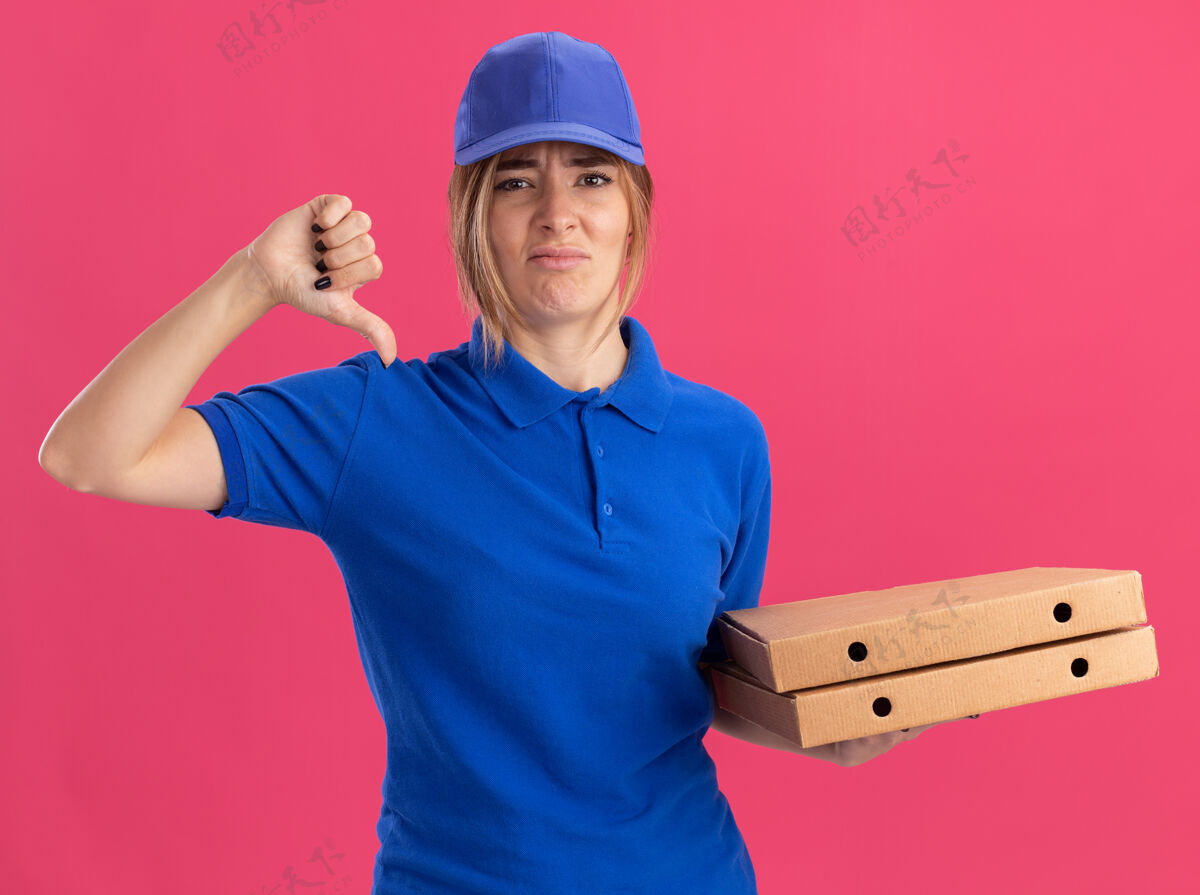 拇指穿着制服的年轻漂亮的送货女孩拿着披萨盒 拇指朝下放在粉红色的披萨上姿势盒子不愉快