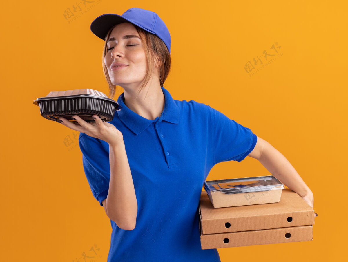 请穿着制服的年轻漂亮的送货女孩高兴地拿着纸食品包 嗅着放在橘色披萨盒上的容器制服嗅食物