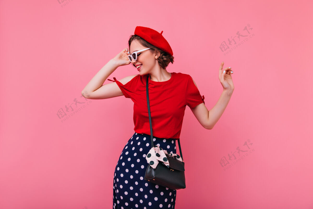 年轻穿着贝雷帽的漂亮时髦女人抚摸着她的太阳镜身着法式服装的快乐卷曲的黑发女孩的室内照片贝雷帽成人欢乐