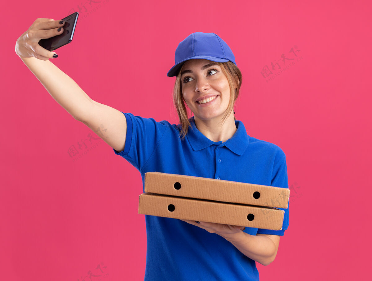 制服身着制服的年轻漂亮的送货女孩微笑着拿着比萨饼盒 看着粉色的手机盒子送货姿势
