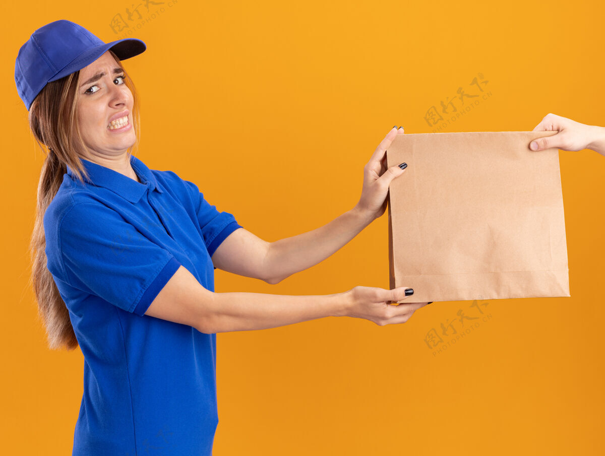 年轻一个穿着制服的年轻漂亮的送货女孩把纸包给了一个看着橙色相机的人制服送货摆姿势