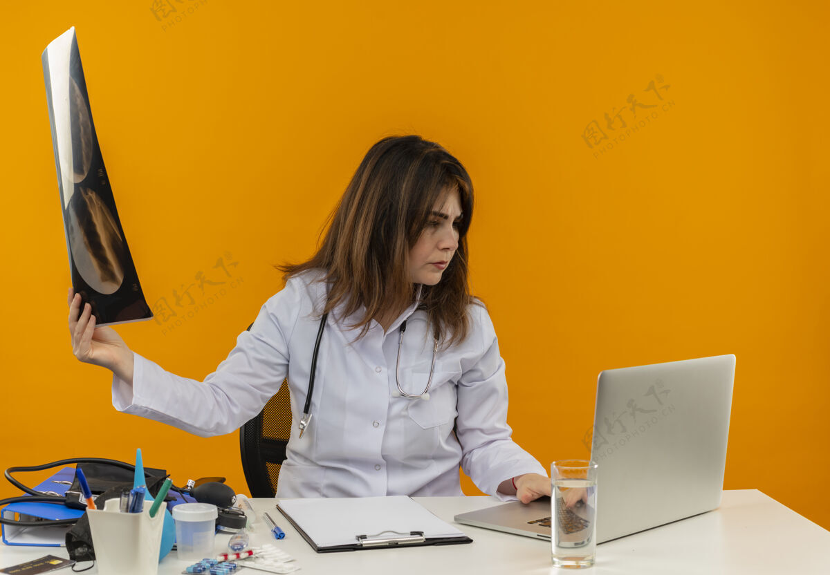 女性集中中年女医生穿着医用长袍和听诊器坐在办公桌前用医用工具夹板拿着x光拍用笔记本电脑隔离X光成人女士
