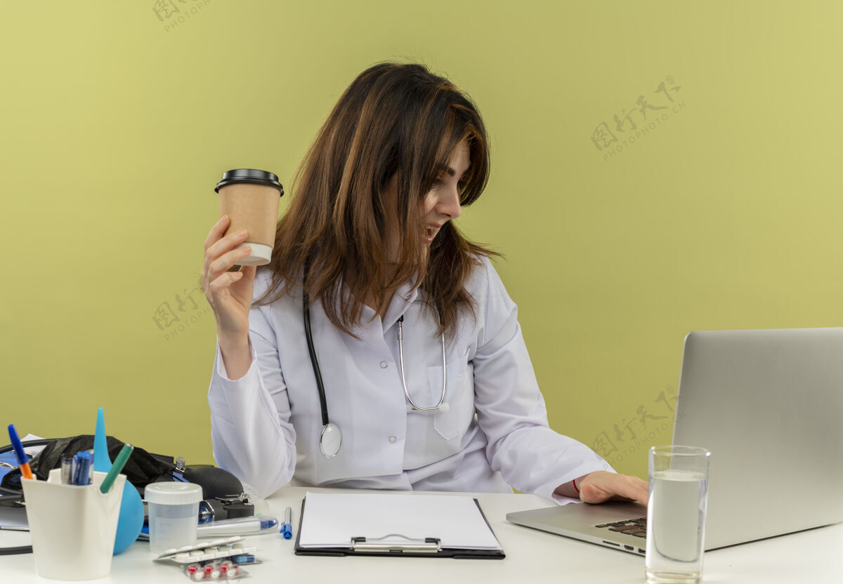 咖啡印象深刻的中年女医生穿着医用长袍和听诊器坐在办公桌旁 用笔记本电脑拿着医用工具和剪贴板 把塑料咖啡杯隔离开坐着女士工具
