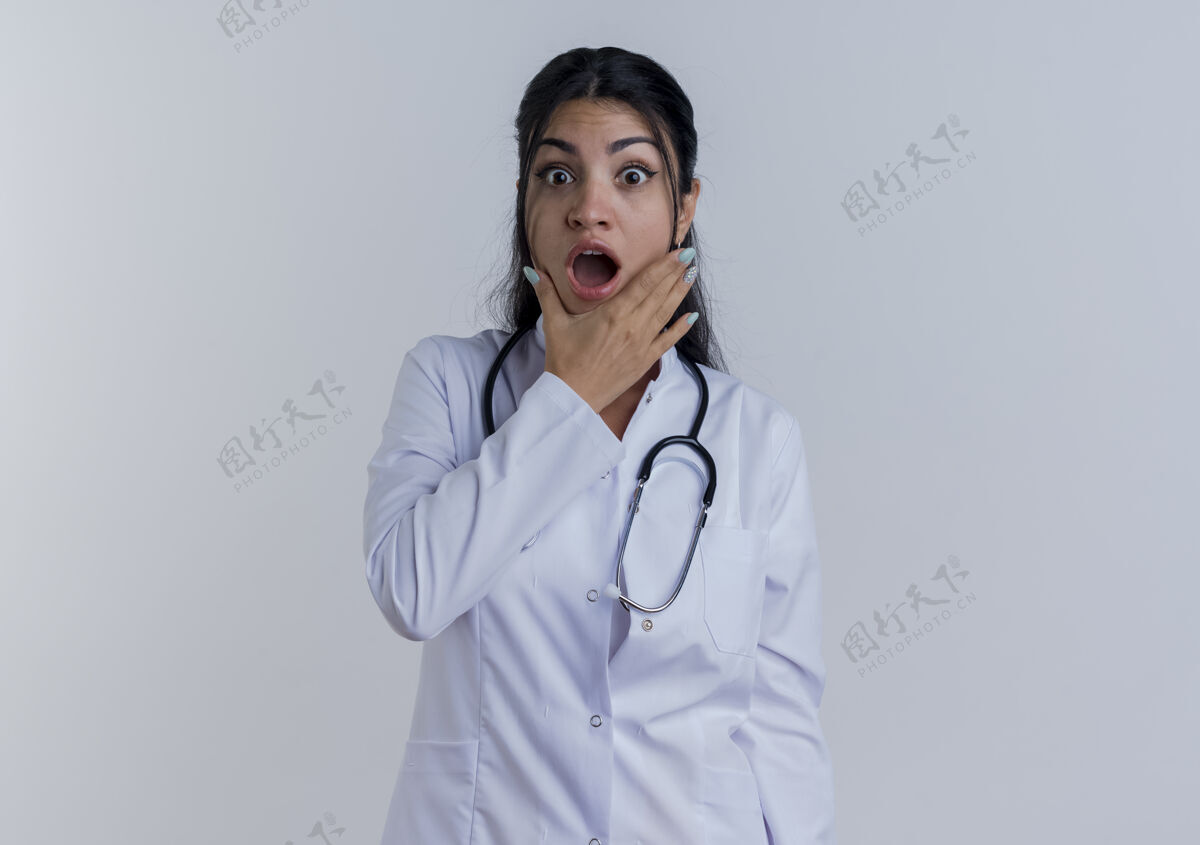 成年人惊讶的年轻女医生穿着医用长袍和听诊器看着把手放在下巴上手女人长袍