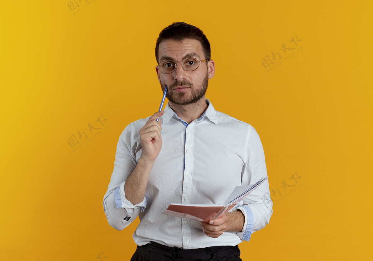 自信自信的帅哥戴着眼镜 把笔放在脸上 把笔记本孤立地放在橙色的墙上持之以恒男人眼镜