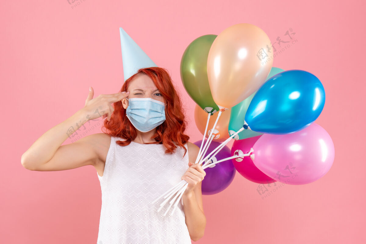 粉色正面图：年轻女性手持彩色气球 戴着无菌面具站在粉红色的椅子上彩色病毒多彩