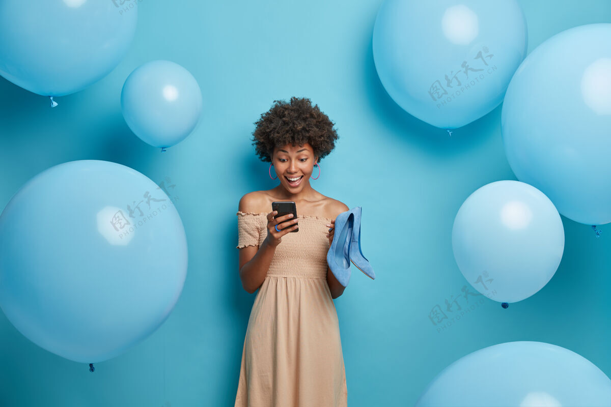 年轻快乐的一天和庆祝的概念积极的深色皮肤的女人在社交网络聊天 穿米色长裙和持有蓝色的鞋子 选择最好的服装看起来辉煌 在蓝色墙上孤立卷发风格鞋