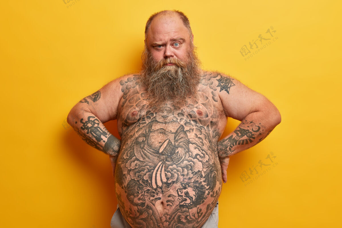 体重大胡子胖子有浓密的胡须 纹身的身体和大肚子 从眉毛下面看 手放在腰上 隔离在黄色的墙壁上肥胖 抽脂 减肥概念男人身材腹部