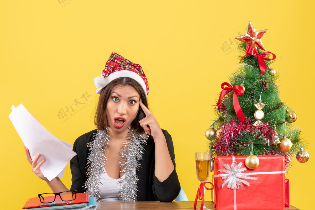 漂亮穿着西装 戴着圣诞老人帽和新年装饰品 手里拿着文件 坐在一张桌子旁 桌上放着圣诞树的办公室里的一位惊讶的商务女士快乐惊喜桌子