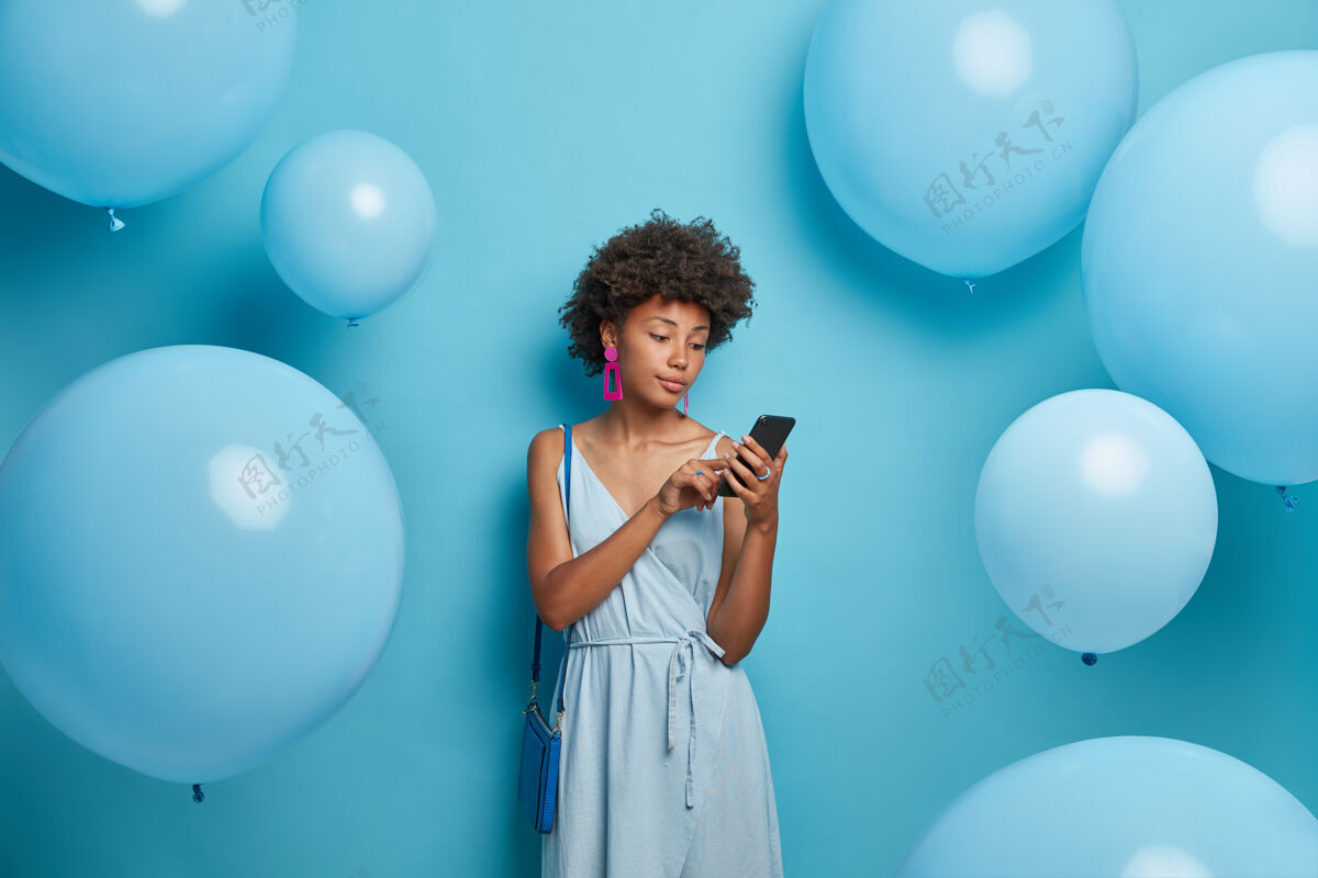 使用优雅的年轻女子在社交网络上聊天 专注于智能手机屏幕 在聚会上感到无聊 尝试新的应用程序 穿着一种颜色的时髦衣服和包 对着蓝色的墙壁摆姿势应用程序女士移动