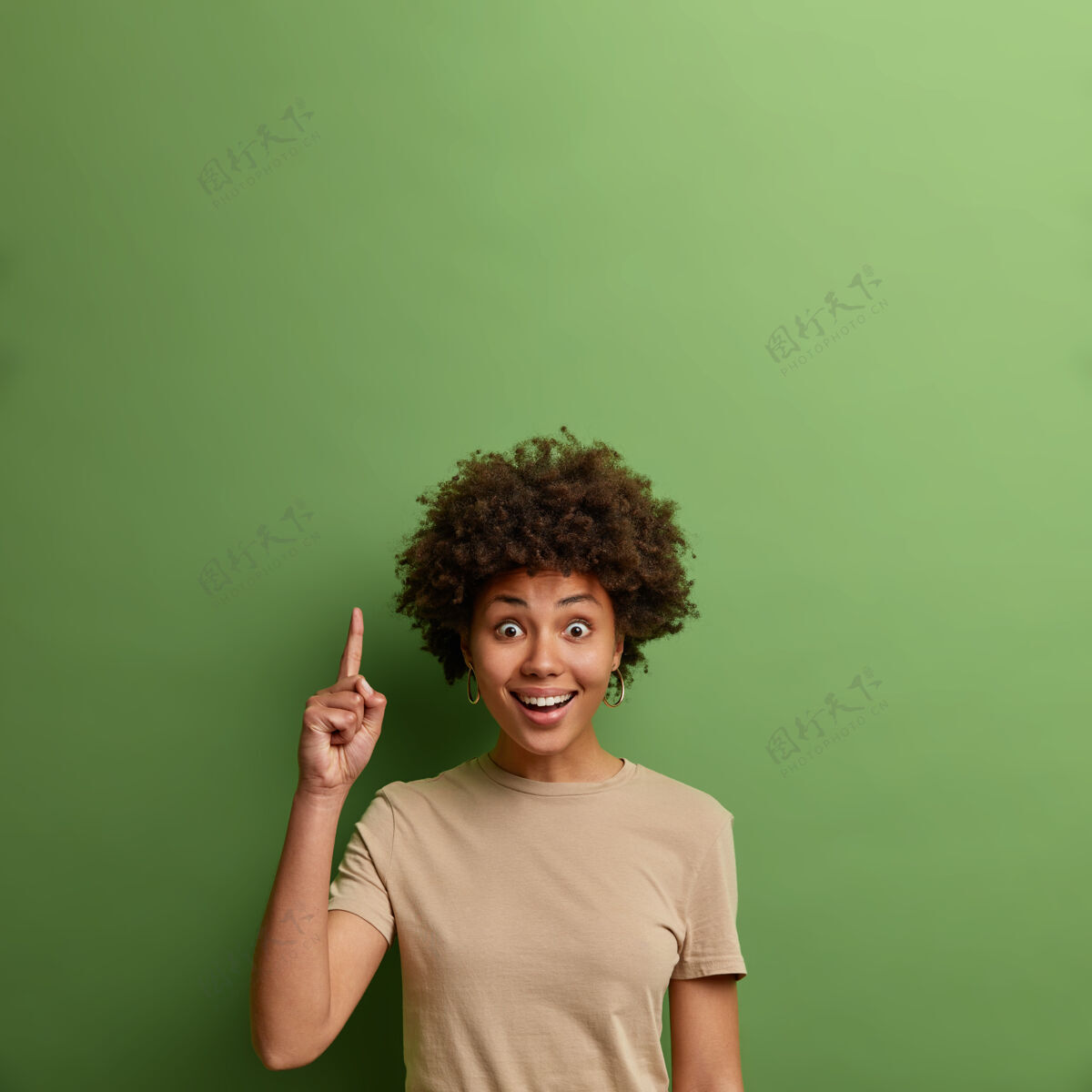 满意快乐惊讶的卷发成年女性指着空白的商业文本 展示创意展示 向上展示精彩的宣传片 穿着休闲米色t恤 绿色墙壁休闲垂直注意
