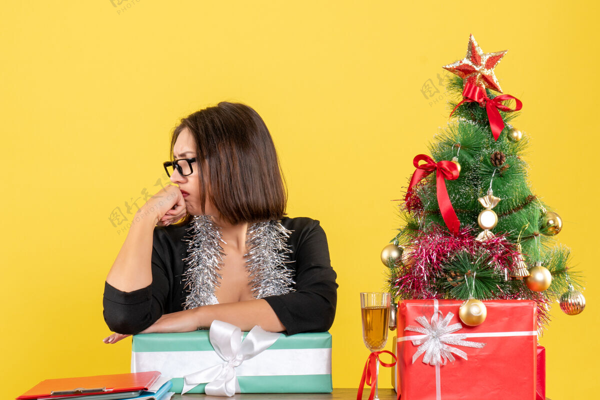 套装一位穿着西装 戴着眼镜展示礼物 坐在一张桌子旁 桌上放着一棵圣诞树的不幸的商务女士坐着微笑树