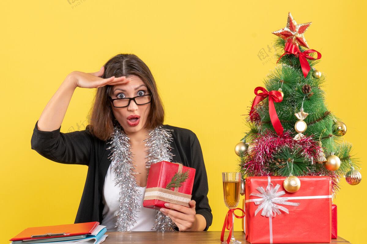 坐着一位穿着西装 戴着眼镜的商务女士拿着礼物 坐在办公室的一张桌子旁 桌上放着一棵圣诞树漂亮人快乐
