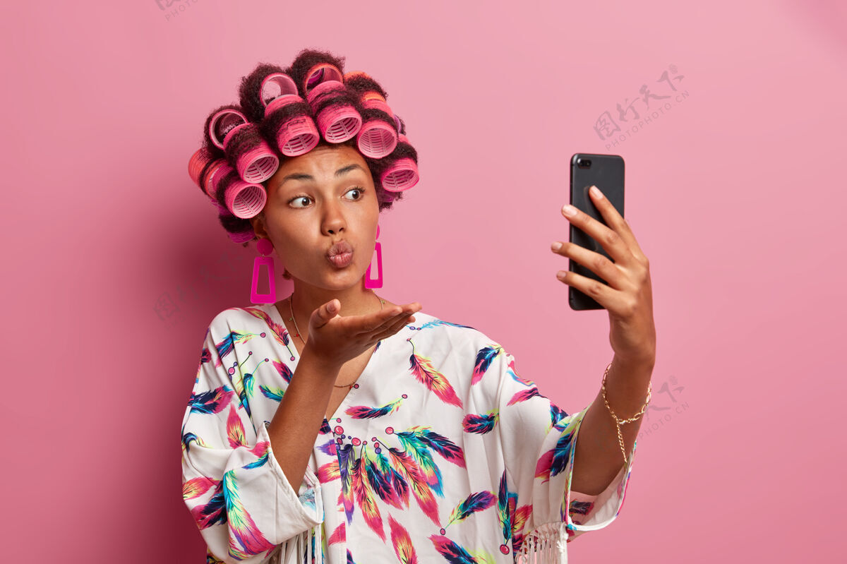 家务淋浴后头上戴着卷发的浪漫美女 通过手机拍摄自拍肖像 吹mwah 穿着休闲家装 享受与男友的视频通话 拥有自然美姿势自拍室内