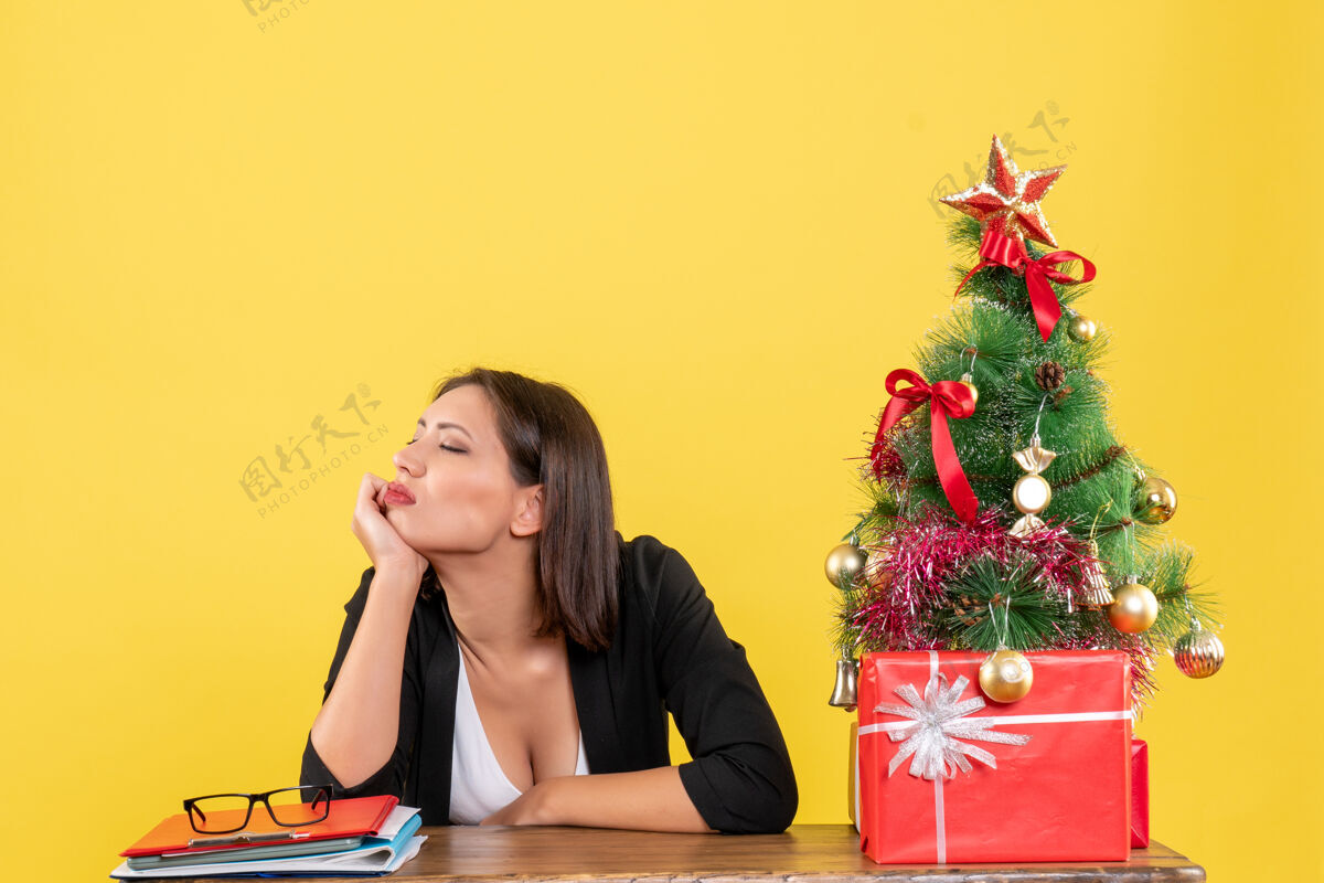 圣诞节不满意的年轻女子在附近的西装装饰圣诞树在黄色的办公室人快乐漂亮
