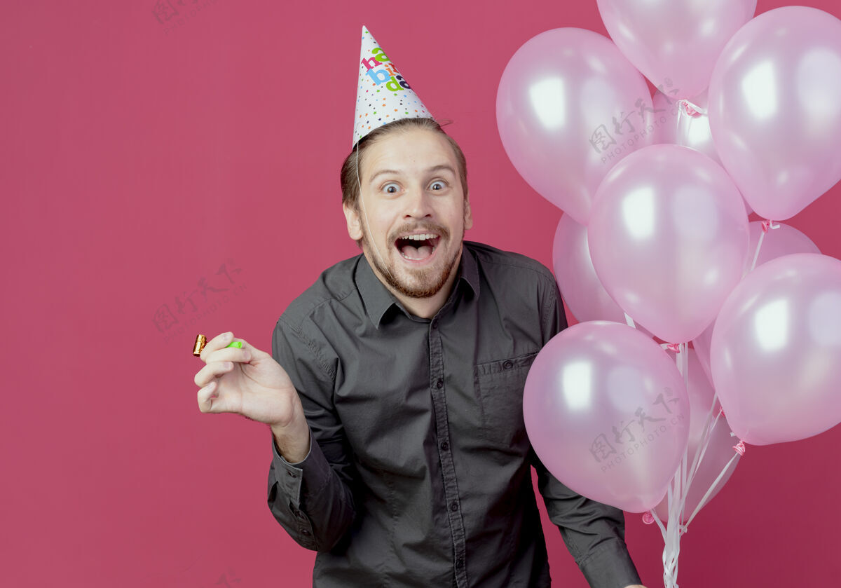 快乐戴着生日帽的快乐帅哥站在粉红色的墙上 手里拿着氦气球 手里拿着哨子粉色男人氦