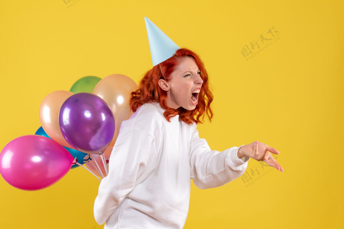 五颜六色正面图年轻女子藏着可爱的彩色气球 背景是黄色的派对新年色彩感人的女人漂亮吹快乐